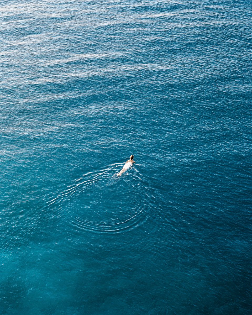 A single swimmer far below on open water, ripples spreading outward.